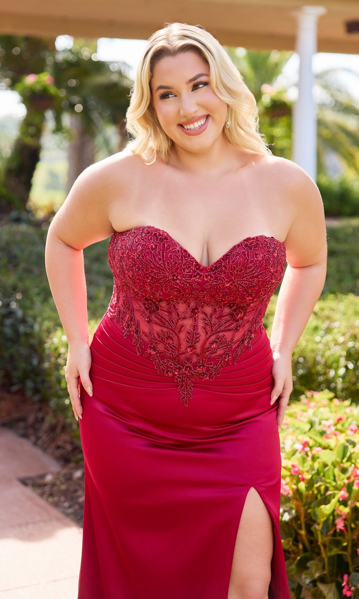 A woman, outdoors and smiling, wears the Long Plus-Size Prom Dress: Primavera 14109—a strapless gown with a sheer embroidered bodice, satin sheath skirt, and thigh-high slit.
